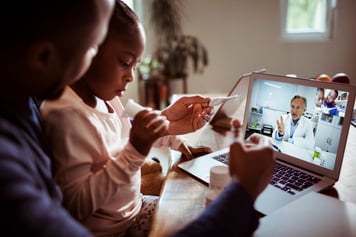 Father using thermometer to take young daughter's tempurature while using laptop for telemedicine video call with docto in white labcoat