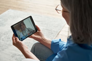 Woman sitting on sofa using tablet for video telemedicine call with female healthcare provider