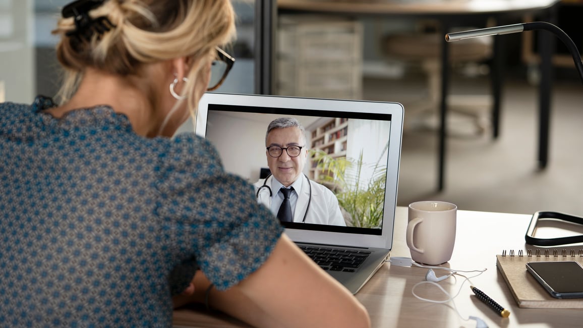 Woman sitting at table using laptop for virtual care call with male doctor