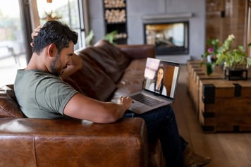 Man sitting on brwn sofa using laptop for video virtual care call with female healthcare provider in labcoat