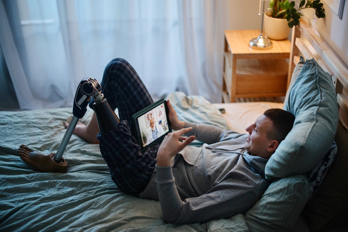 Manle adult amputee laying in bed using tablet for video telemedicine call with male doctor in labcoat