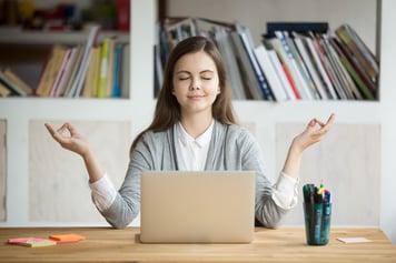 Young adult woman sitting at table meditating with laptop open in front of her